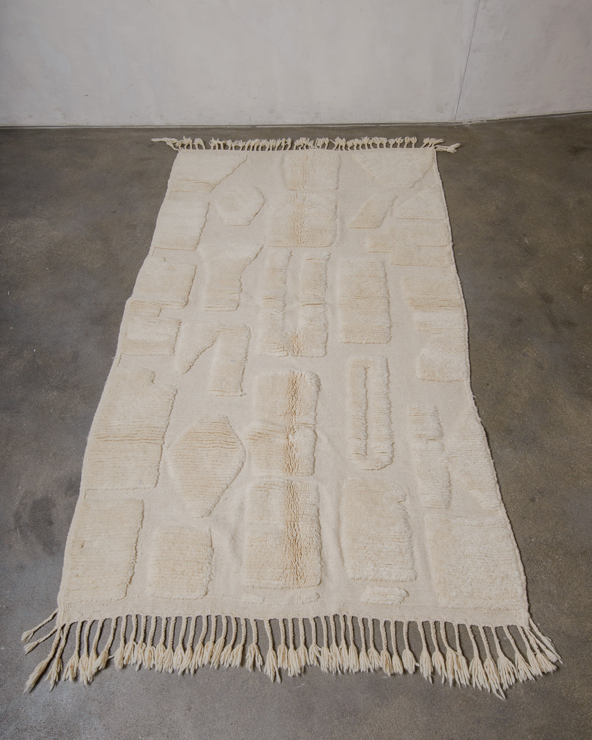 Beige woven wool rug with geometric forms and fringes on a concrete floor.